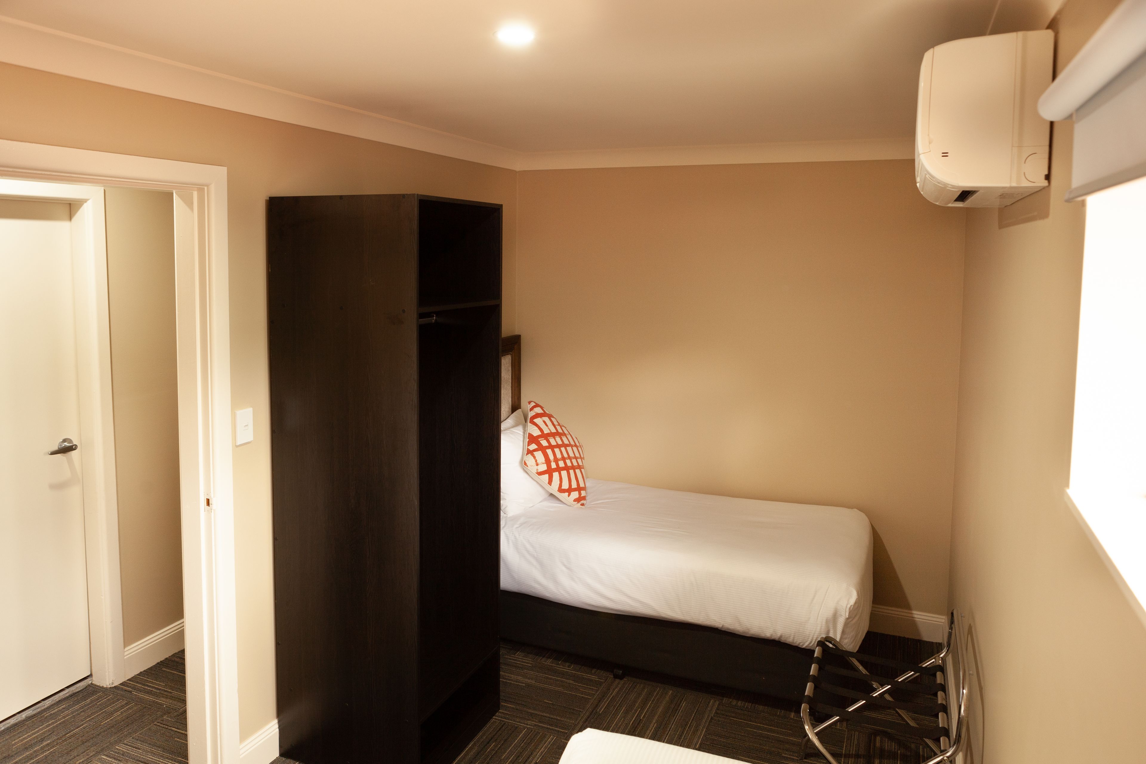 A small, single-bed room with a dark wardrobe, a white wall-mounted air conditioner, and a window providing natural light.