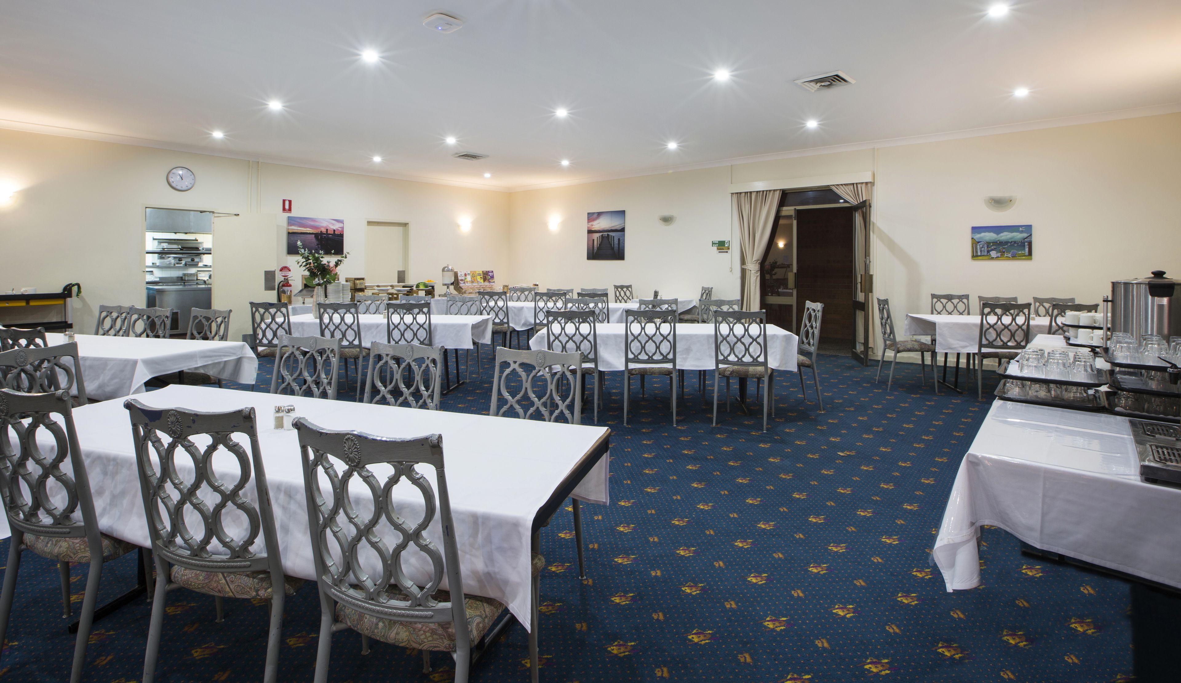 A spacious dining area with multiple tables and chairs, a kitchen in the background, and a clock on the wall.