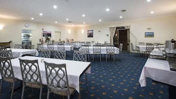 A spacious dining area with multiple tables and chairs, a kitchen in the background, and a clock on the wall.