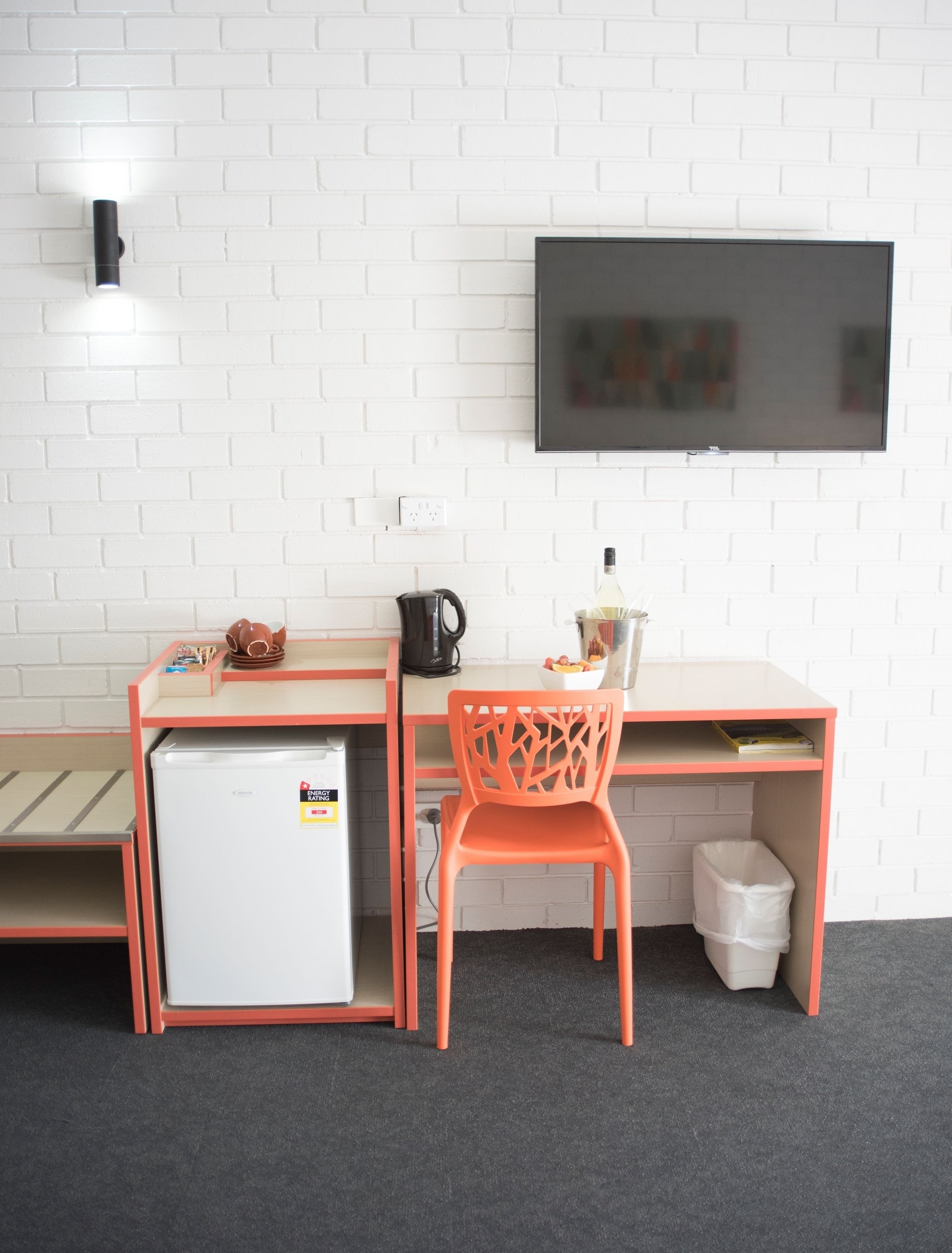A compact room with a kitchenette, a single orange chair, a small white fridge, a TV mounted on the wall, and a trash bin.