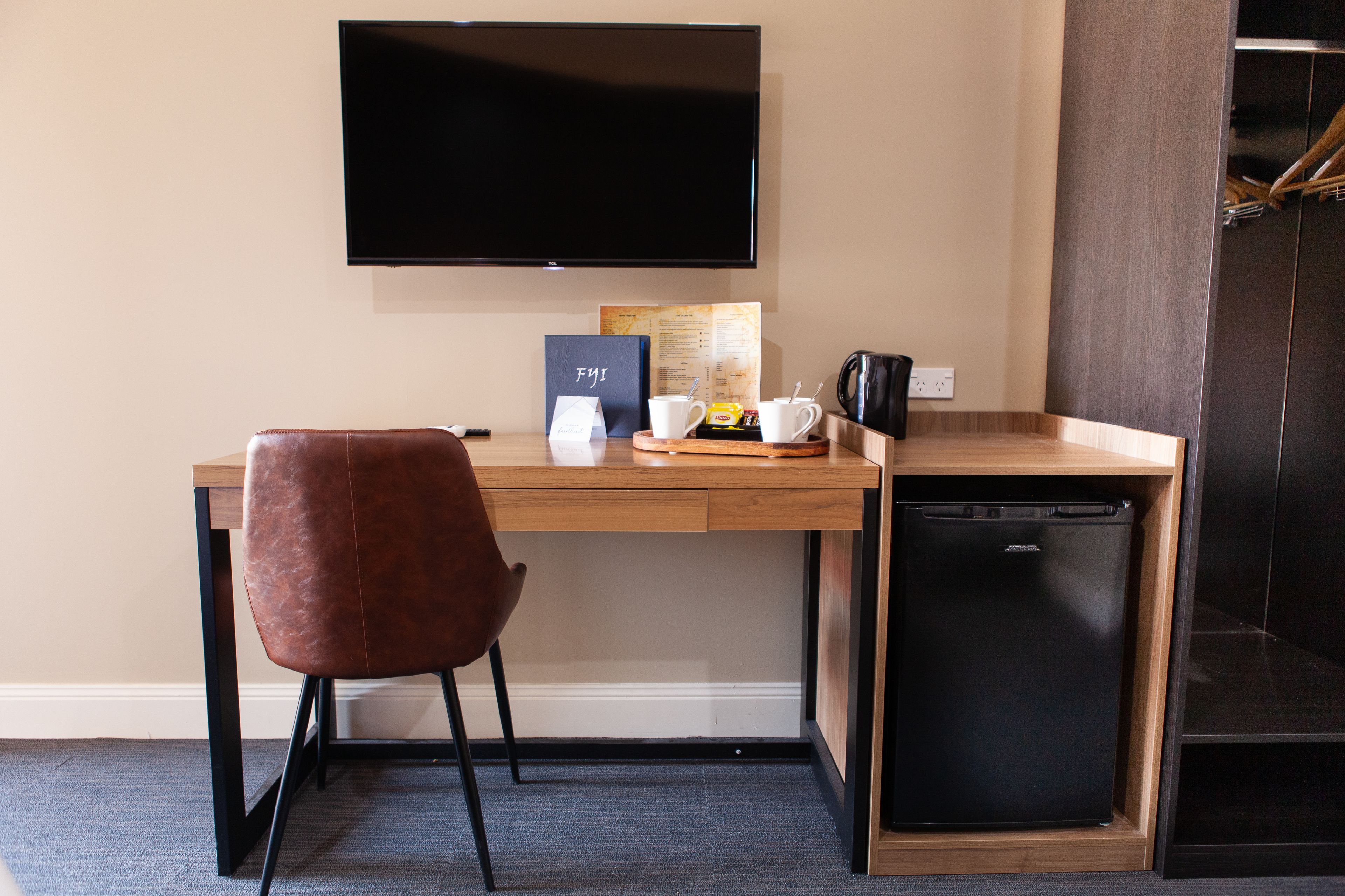 Quarto de hotel compacto com escrivaninha de madeira, poltrona de couro marrom, pequeno frigorífico preto, máquina de café e televisão de ecrã plano fixada na parede.
