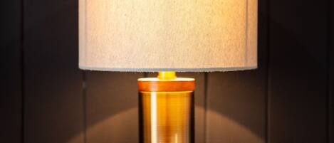 A table lamp with a cylindrical base and a fabric shade, placed on a wooden surface against a dark background.