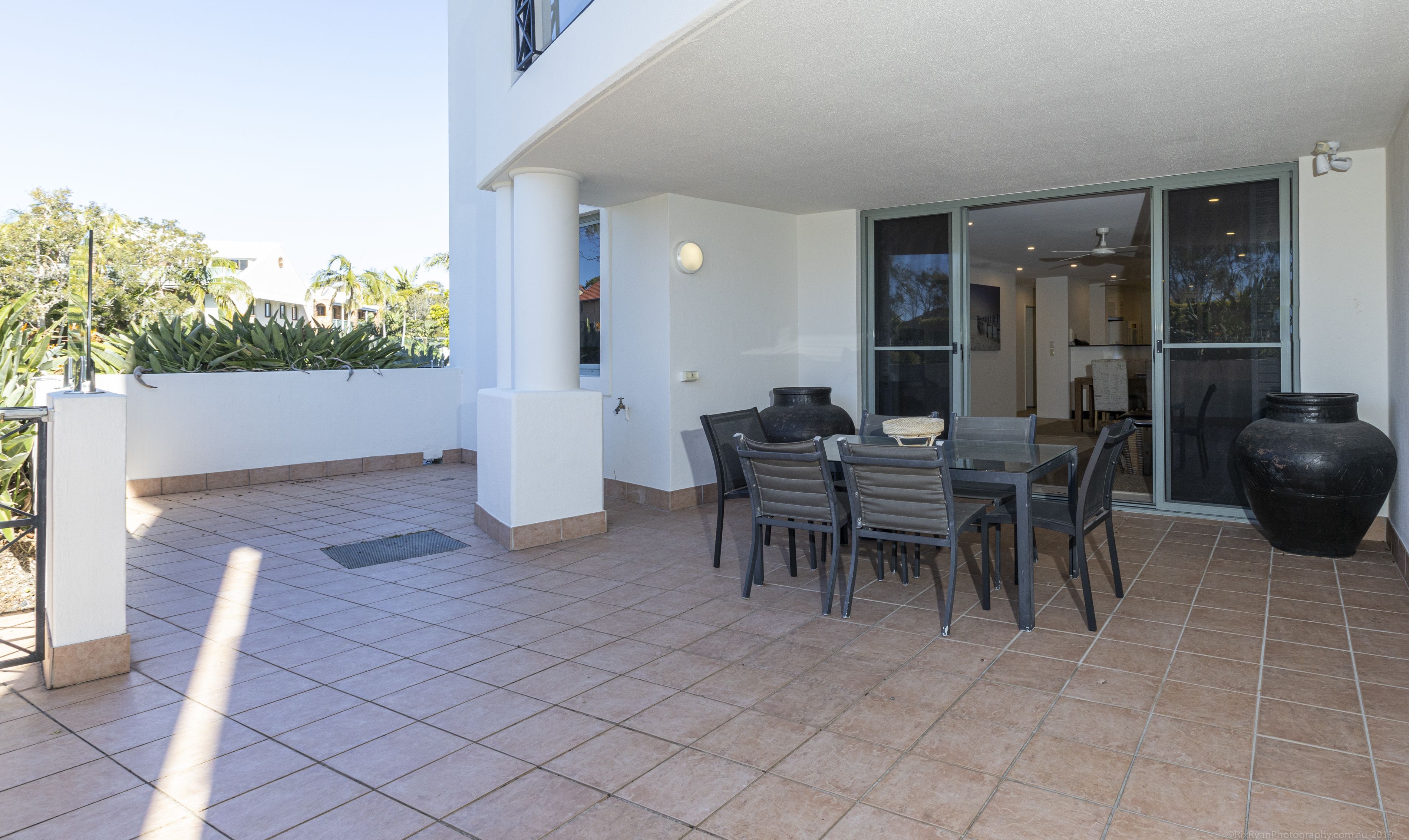 Une terrasse avec une table et des chaises, un grand vase et une porte-fenêtre coulissante donnant sur un espace intérieur.