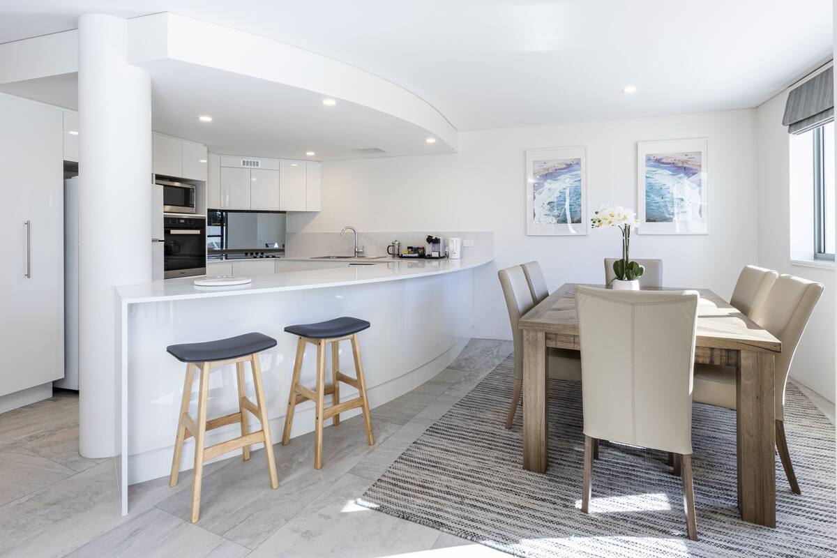 A modern kitchen with white cabinetry, a central island with stools, and a dining area with a wooden table and chairs.