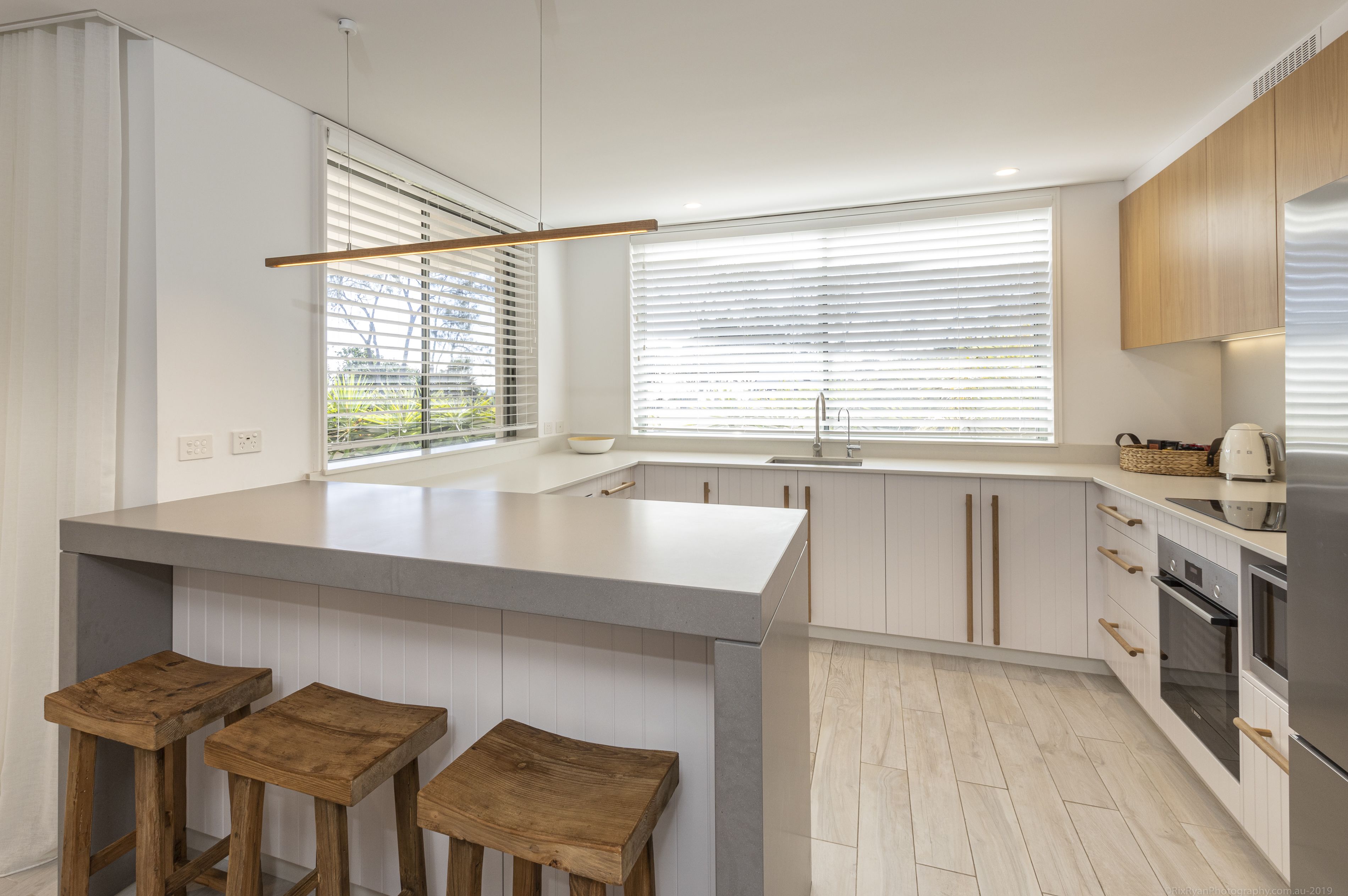 A modern kitchen with a large island, wooden stools, a built-in oven, and a window with blinds.