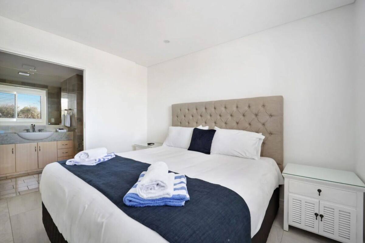 A hotel room with a large bed, a bedside table, a nightstand, a bathroom with a sink and mirror, and a window with blinds.
