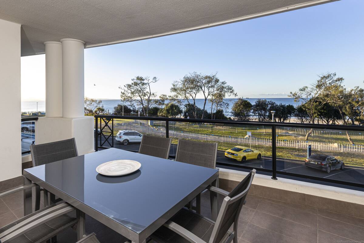 A balcony with a table and chairs, overlooking a parking area and a beach.