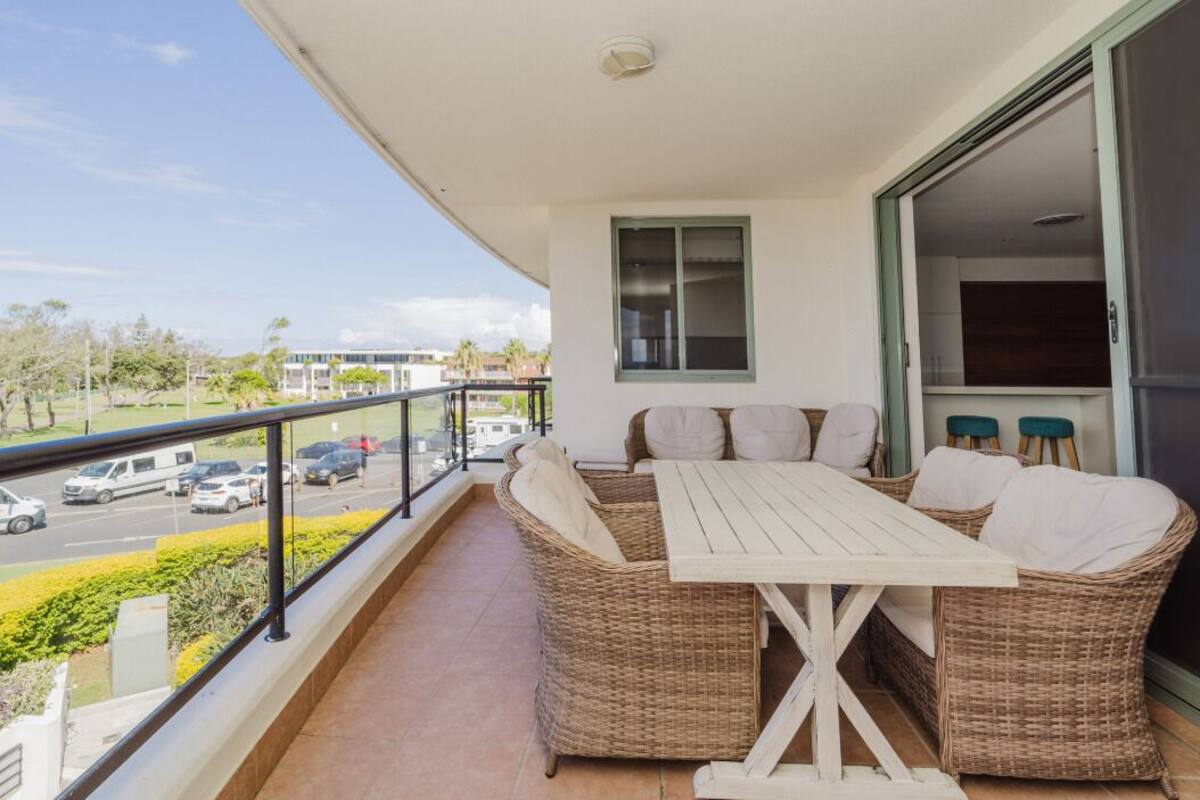 A balcony with a wicker sofa and table, a view of the street, and a door leading to an interior space.