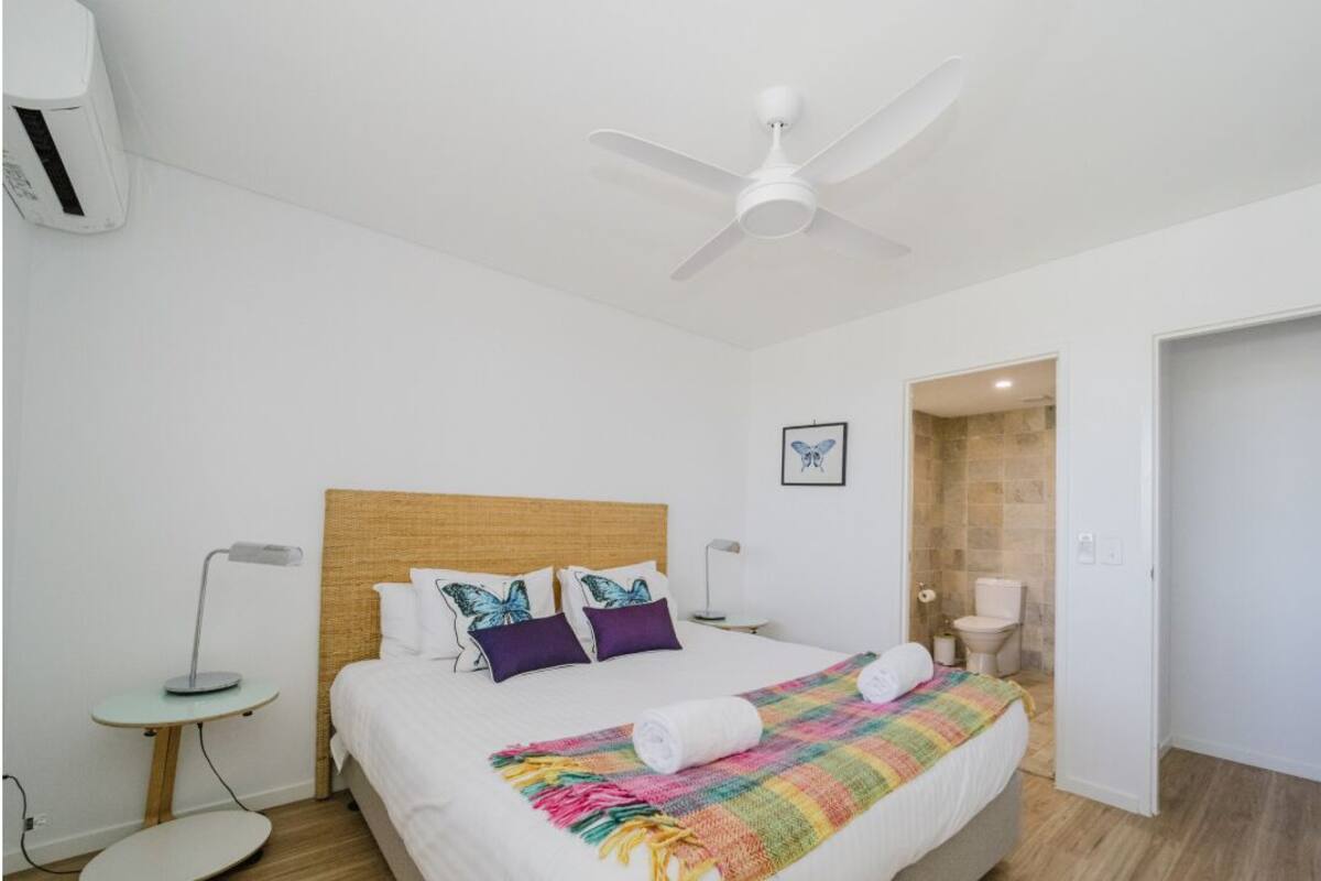 A hotel room with a bed, a ceiling fan, a bedside table with a lamp, and a bathroom visible through an open door.