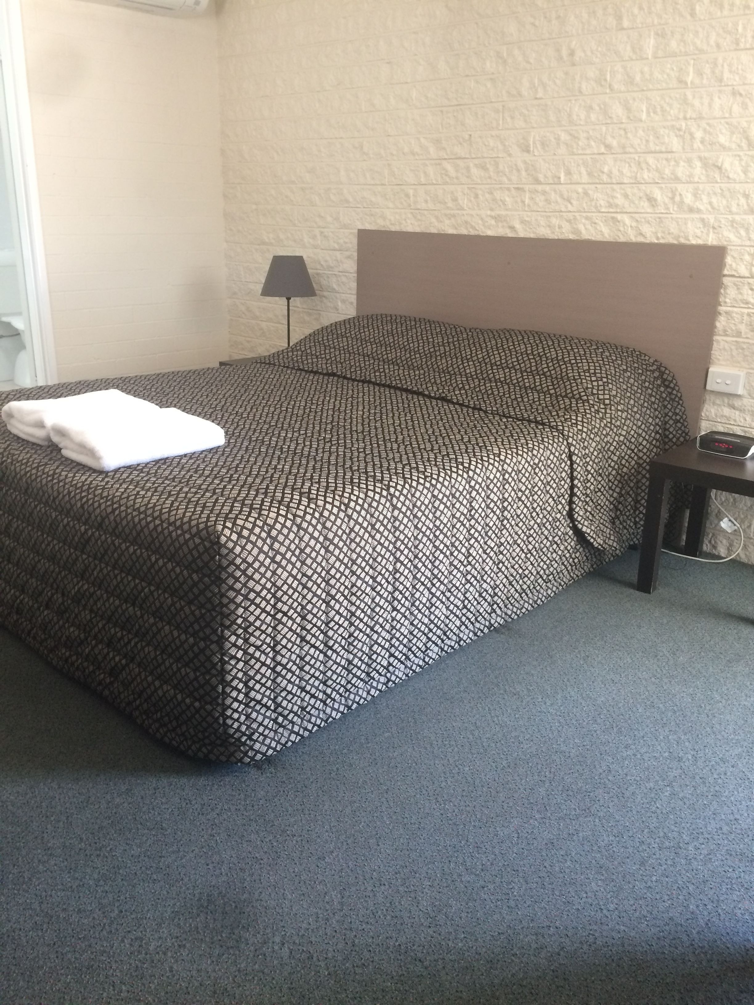 A neatly made bed with a patterned bedspread, a bedside table with a lamp, and a plain wall.