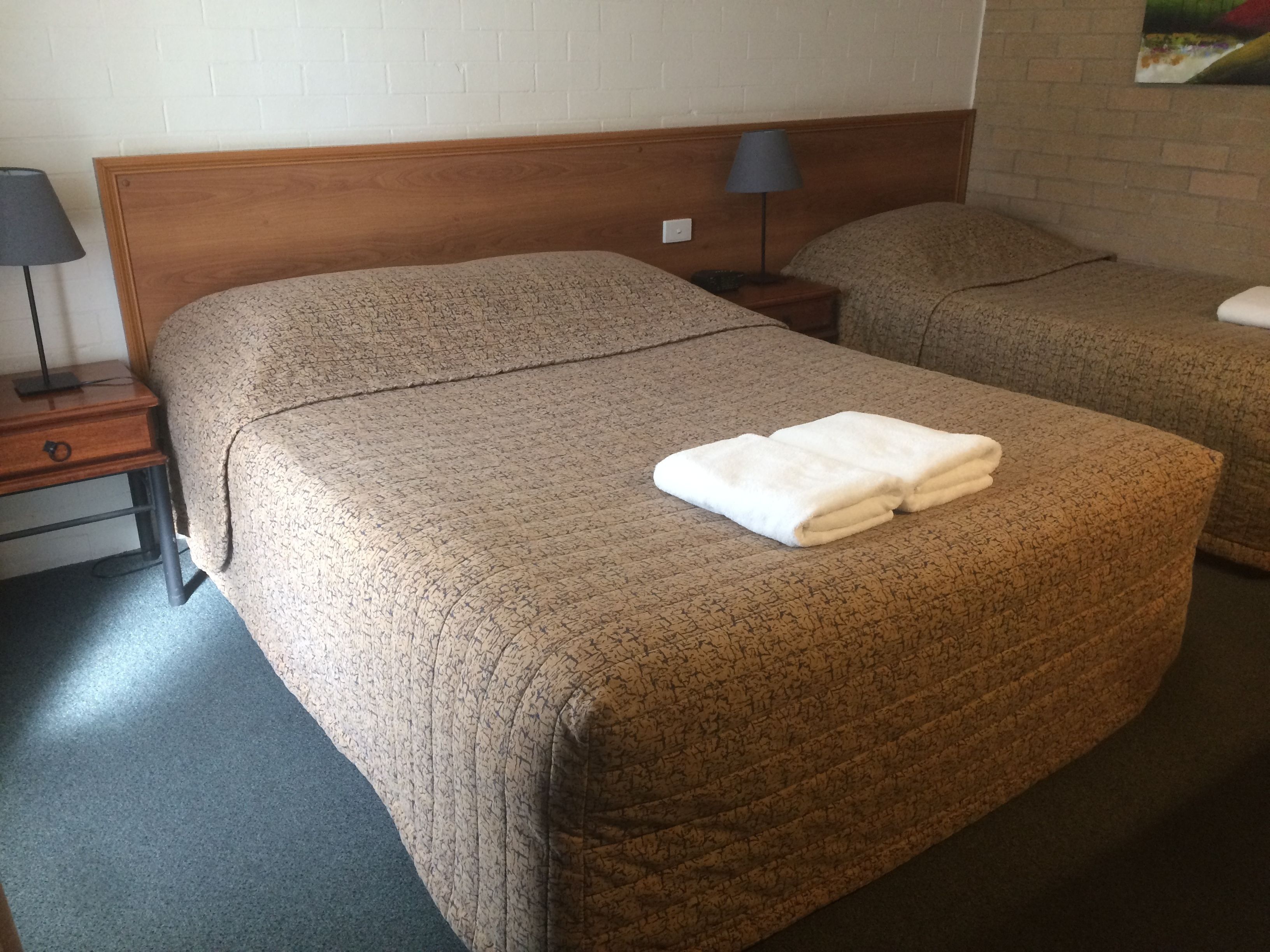 Ein Hotelzimmer mit einem hölzernen Kopfteil, einem Bett mit einem strukturierten, beige farbenen Bettbezug, einem Nachttisch mit einer Lampe und einem weiteren Bett im Hintergrund.