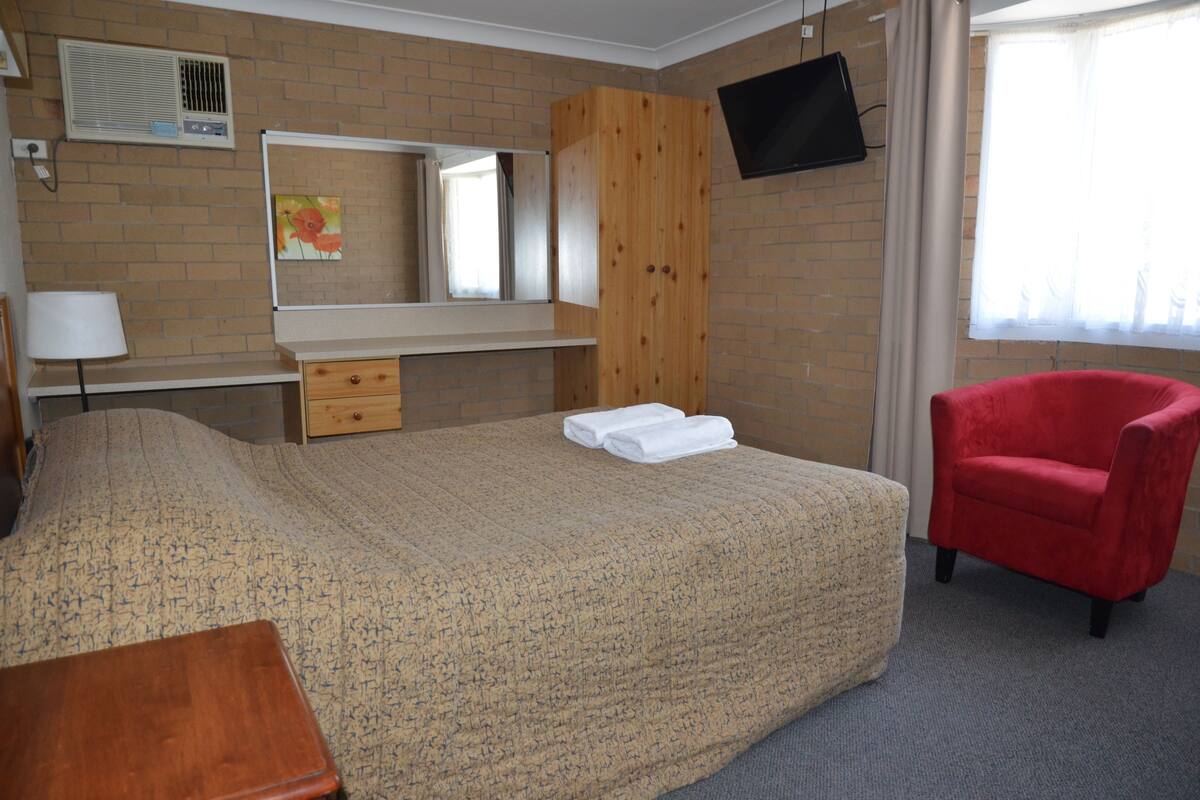 A hotel room with a bed, a wooden wardrobe, a television mounted on the wall, a red chair, and a window with curtains.
