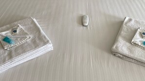 A neatly made bed with white linens and pillows, a remote control, and two towels with toiletries on a textured wall background.