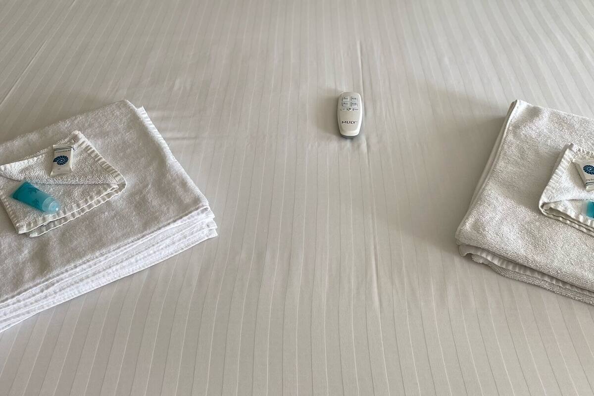 A neatly made bed with white linens and pillows, a remote control, and two towels with toiletries on a textured wall background.