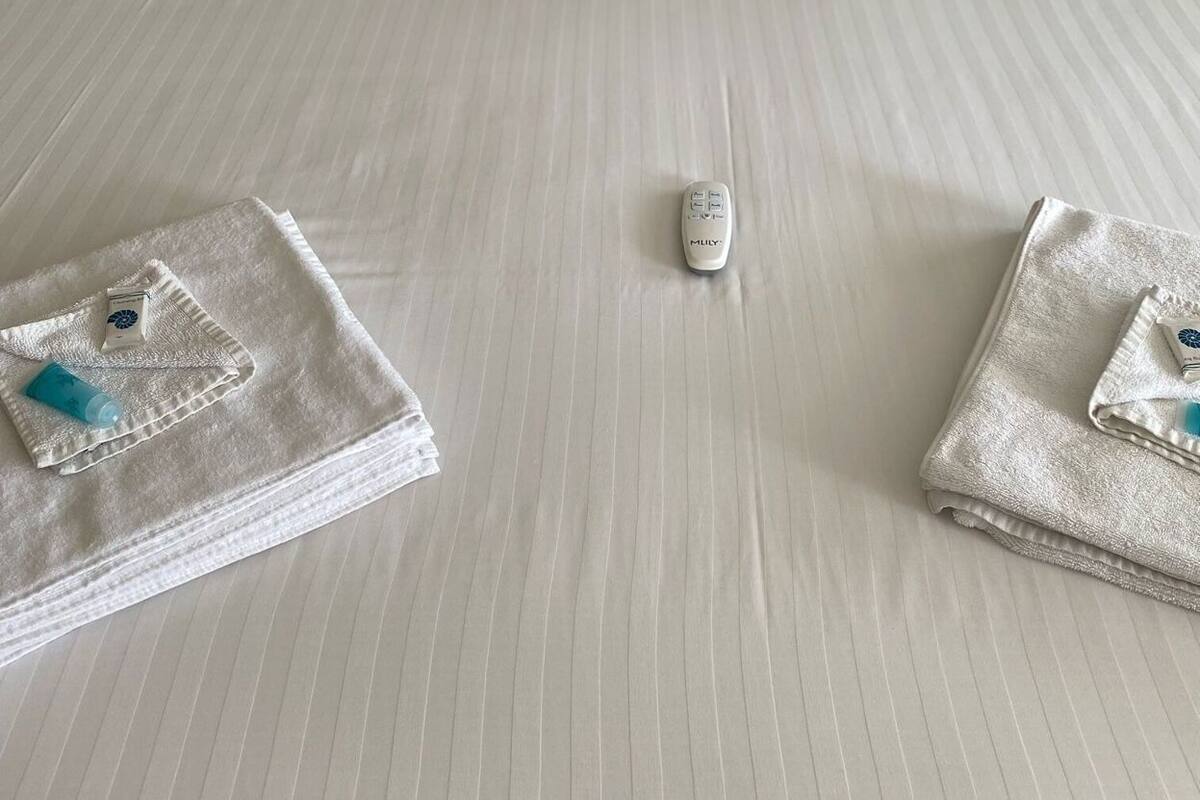 A neatly made bed with white linens, two folded towels, and a remote control on the bed.