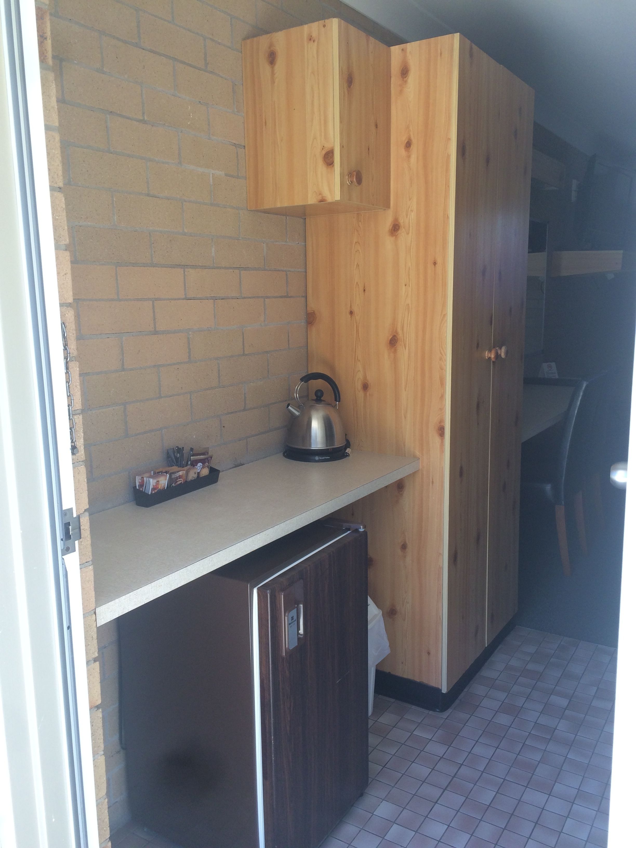 A compact kitchen with wooden cabinets, a stainless steel kettle, and a small refrigerator.
