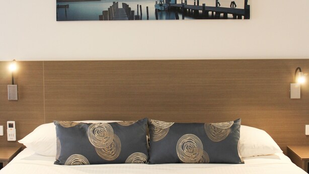 A hotel room with a bed, a headboard, a wall-mounted painting of a cityscape, and two bedside lamps. - Aston Motel Yamba (Yamba)