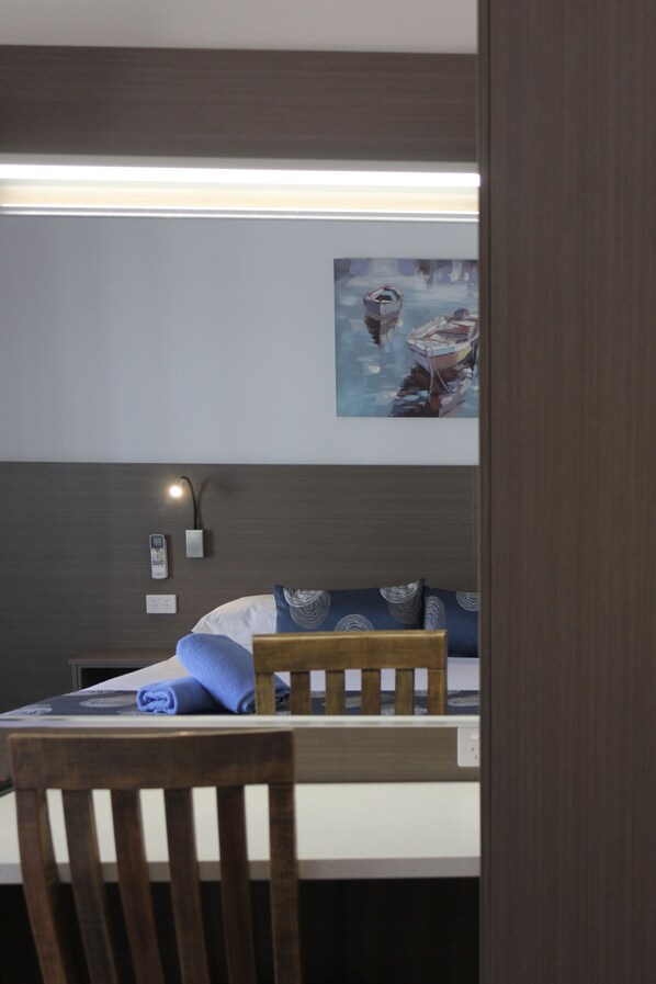 A neatly arranged bedroom with a bed, a bedside lamp, a wall-mounted power outlet, and a painting of boats on the wall.