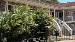 A pool area with a clear blue pool, a staircase leading to a balcony, and lush green palm trees. - Aston Motel Yamba (Yamba)