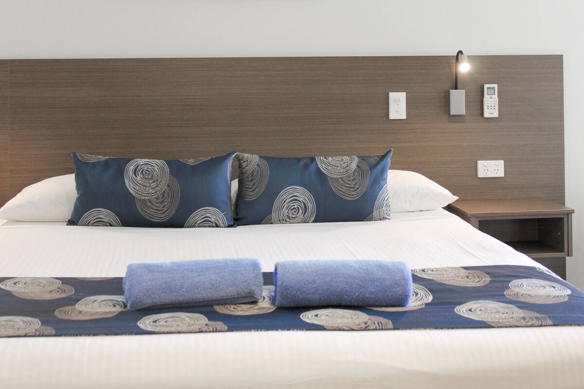 Una cama bien tendida con ropa de cama azul y blanca, un cabecero, lámparas de noche y un enchufe en la mesita de noche.