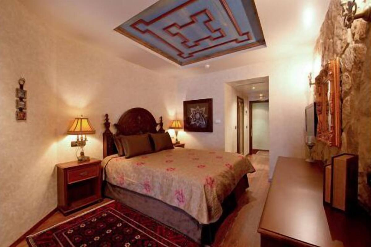 A bedroom with a large bed, two bedside tables, a nightstand, a wooden dresser, a patterned rug, and a ceiling with a geometric design.