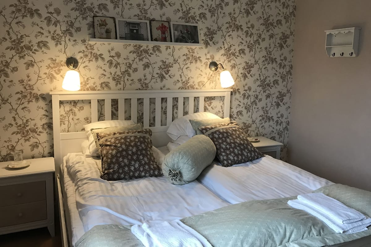 A neatly made bed with floral-patterned pillows and a white headboard. There are two bedside tables with lamps, and a wall-mounted shelf above the bed.