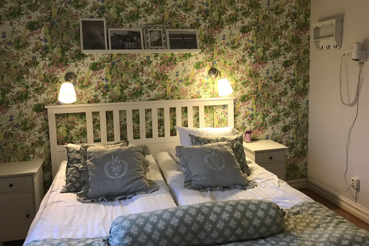 A bedroom with a floral wallpaper, a white headboard, two bedside lamps, and a bed with patterned bedding.