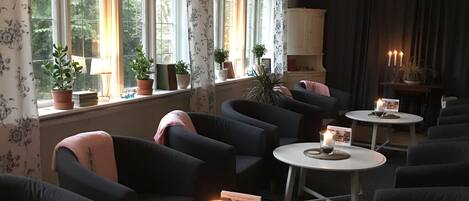Un rincón de descanso acogedor con diversas opciones de asientos, incluyendo sofás y sillones, dispuestos alrededor de pequeñas mesas redondas. Hay plantas en macetas sobre los alféizares de las ventanas y una chimenea con velas.