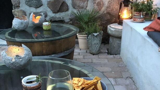 A glass table with a wine glass and chips, surrounded by stone walls and potted plants.