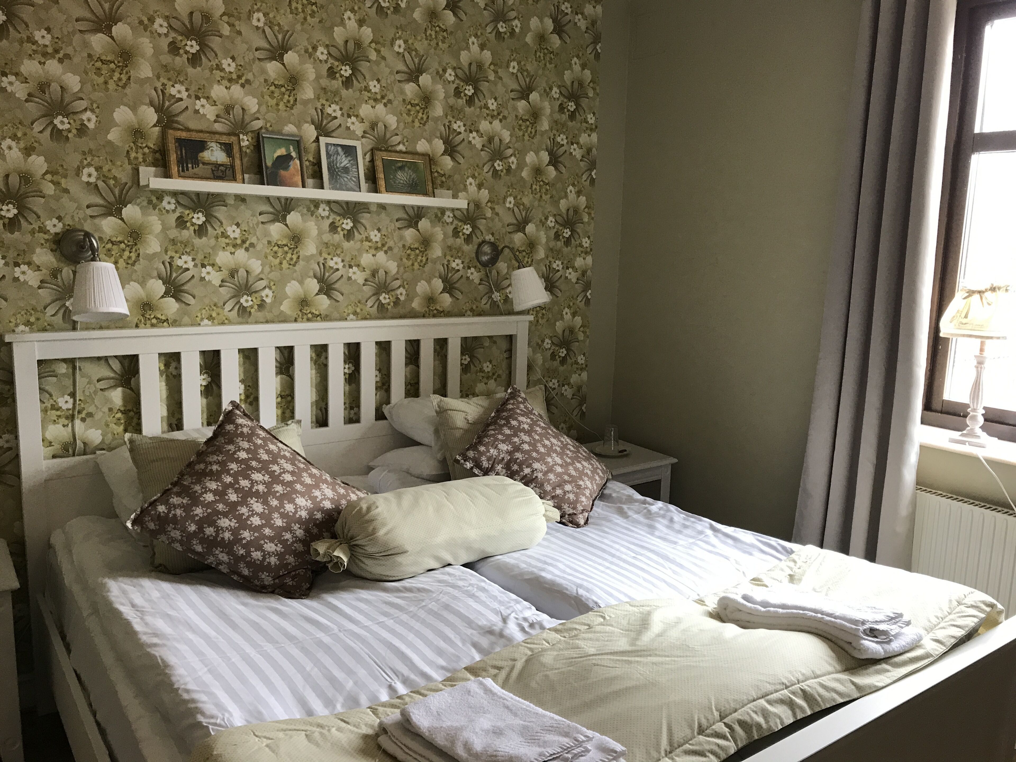 A neatly made bed with striped bedding and floral-patterned pillows, a white headboard, and a small bedside table with a lamp.