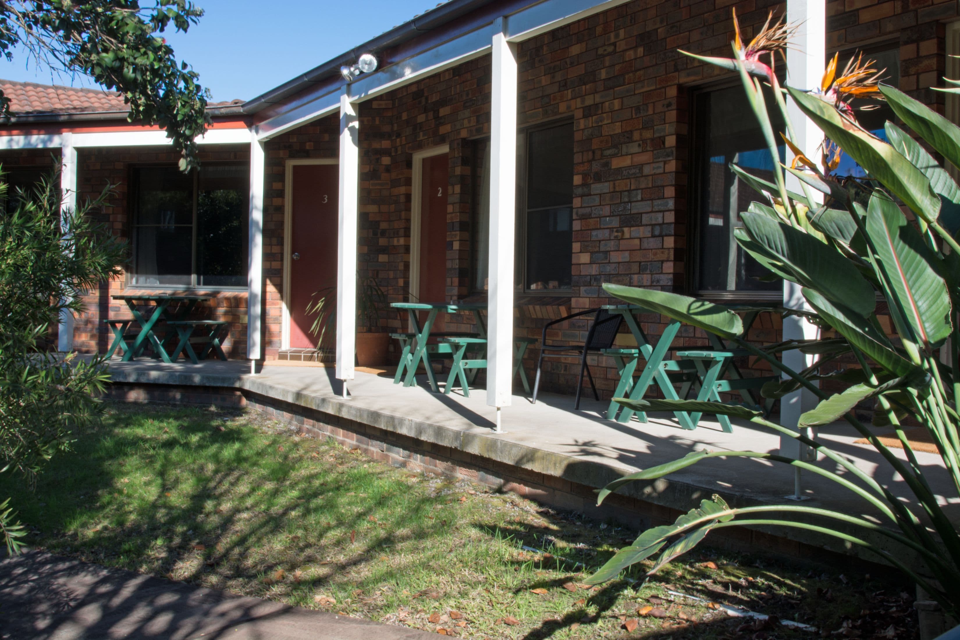 A brick building with a covered patio area, outdoor seating, and a garden with tropical plants.