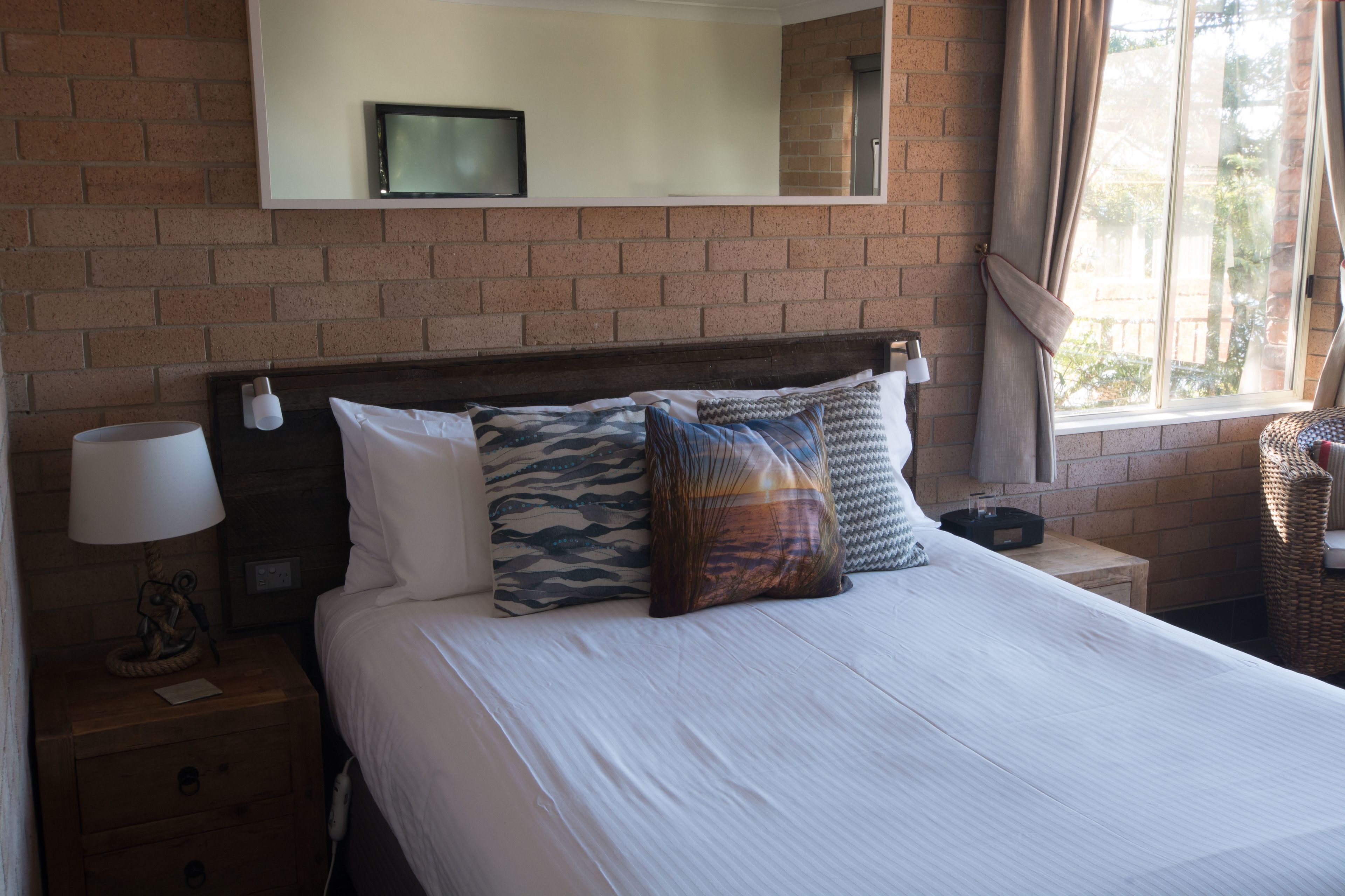 A hotel room with a brick wall, a bed with white bedding and patterned pillows, a bedside lamp, a small table with a lamp, and a window with curtains.