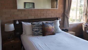 A hotel room with a brick wall, a bed with white bedding and patterned pillows, a bedside lamp, a small table with a lamp, and a window with curtains.