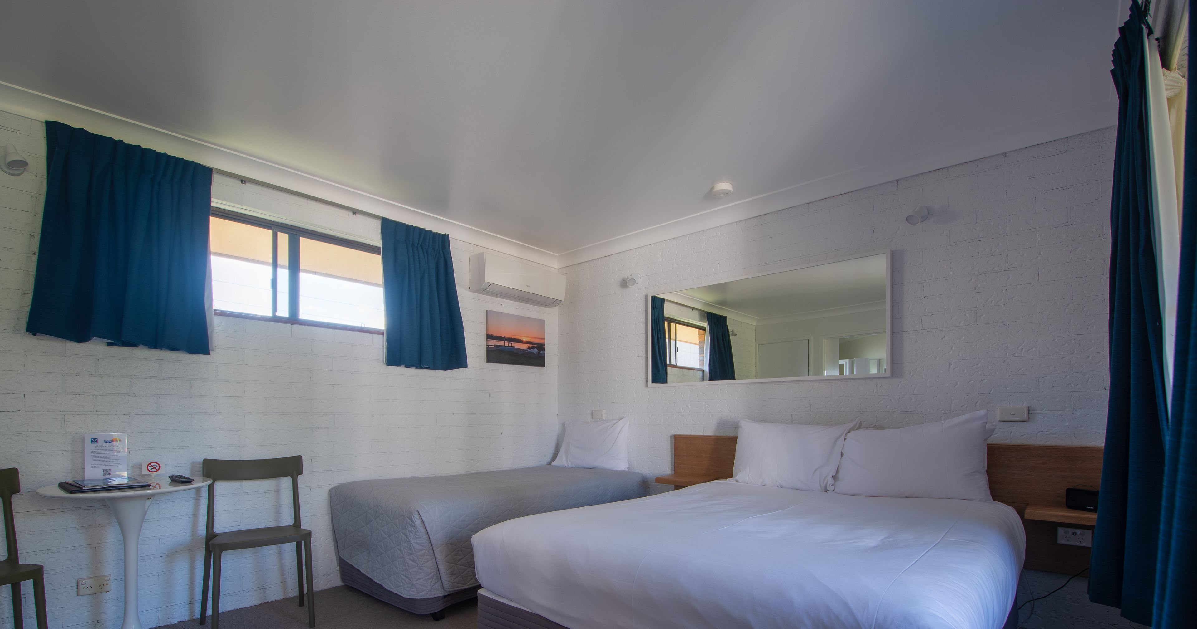 A room with two single beds, a small table with a chair, a mirror, and a window with curtains.