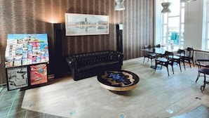 A modern living room with a black sofa, a coffee table, and a wall-mounted TV. There is a dining area with a table and chairs, and a large window providing natural light. - Alpha Hotel (Ostend)