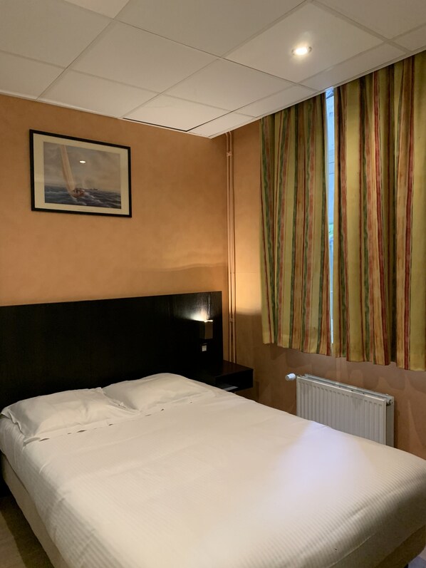 Una habitación de hotel con una cama, una mesita de noche, una lámpara de pared, un cuadro enmarcado y una ventana con cortinas a rayas.