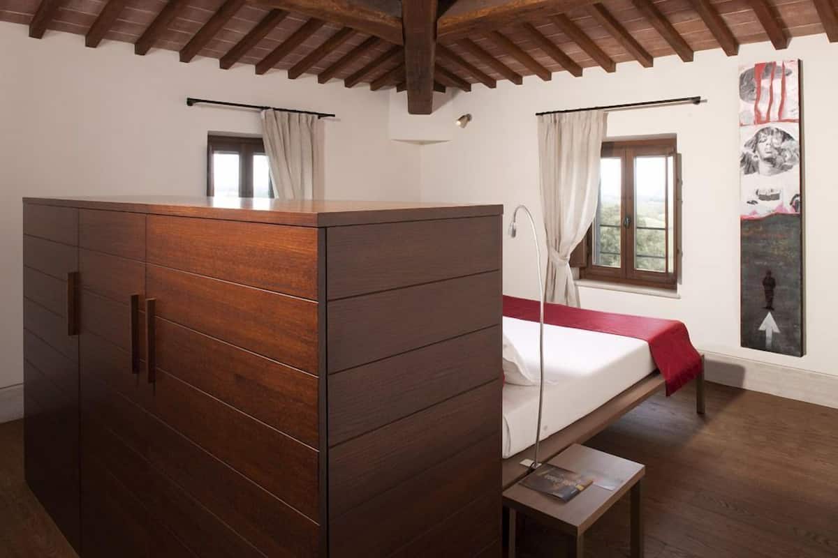 A bedroom with a wooden wardrobe, a bed with a red blanket, a window with curtains, and a wall with artwork.