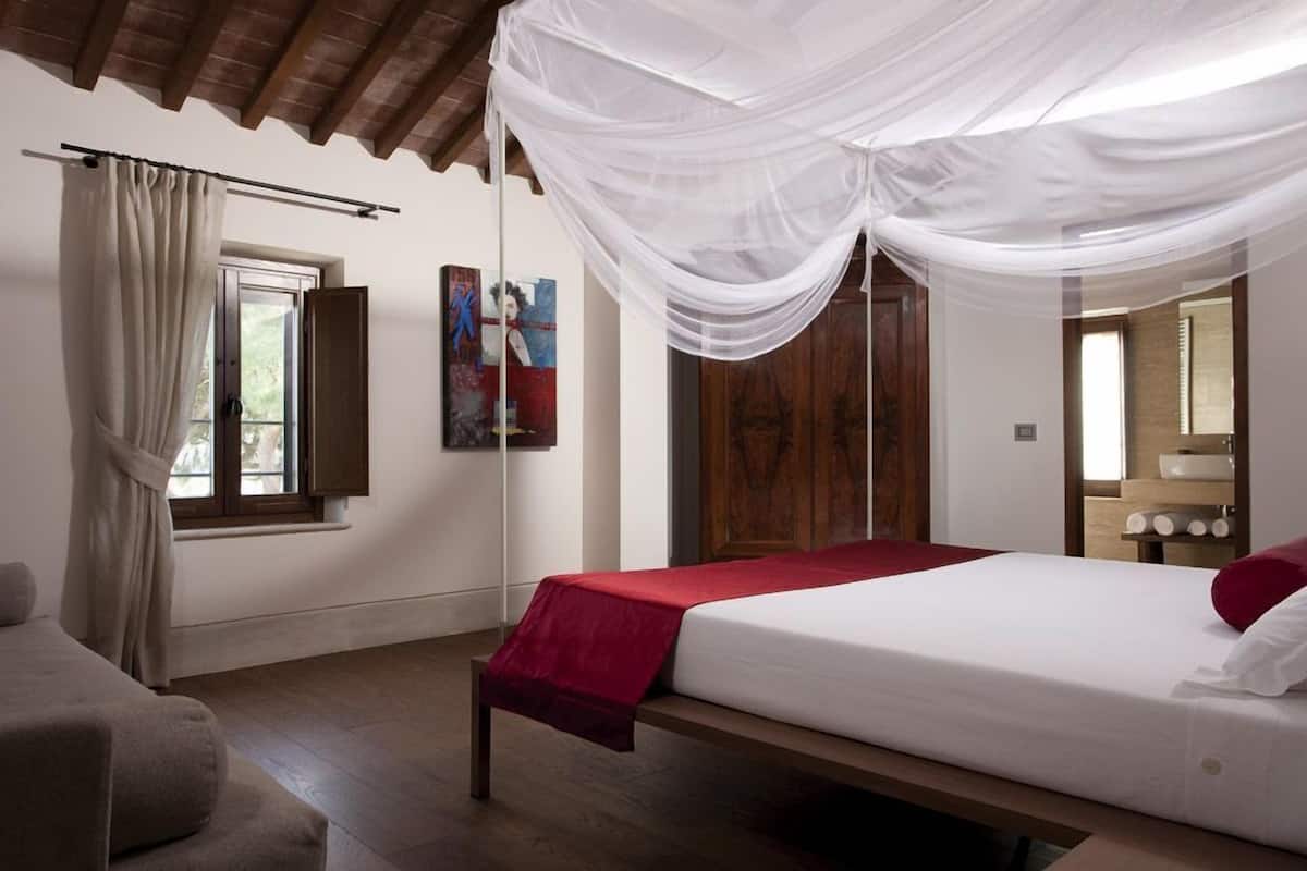 A bedroom with a bed, a wooden bench, a window with curtains, a wooden wardrobe, and a painting on the wall.
