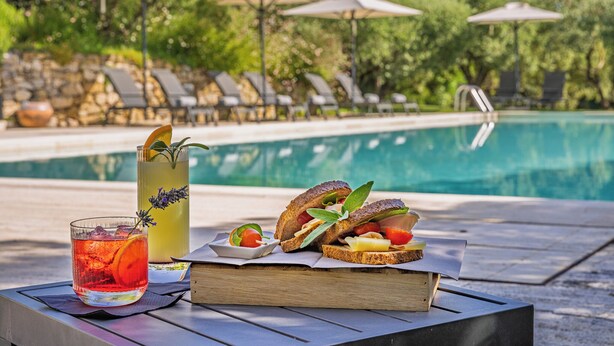 Escena junto a la piscina con una mesa que tiene una bebida, un sándwich y un libro, rodeada de sillones y sombrillas.
