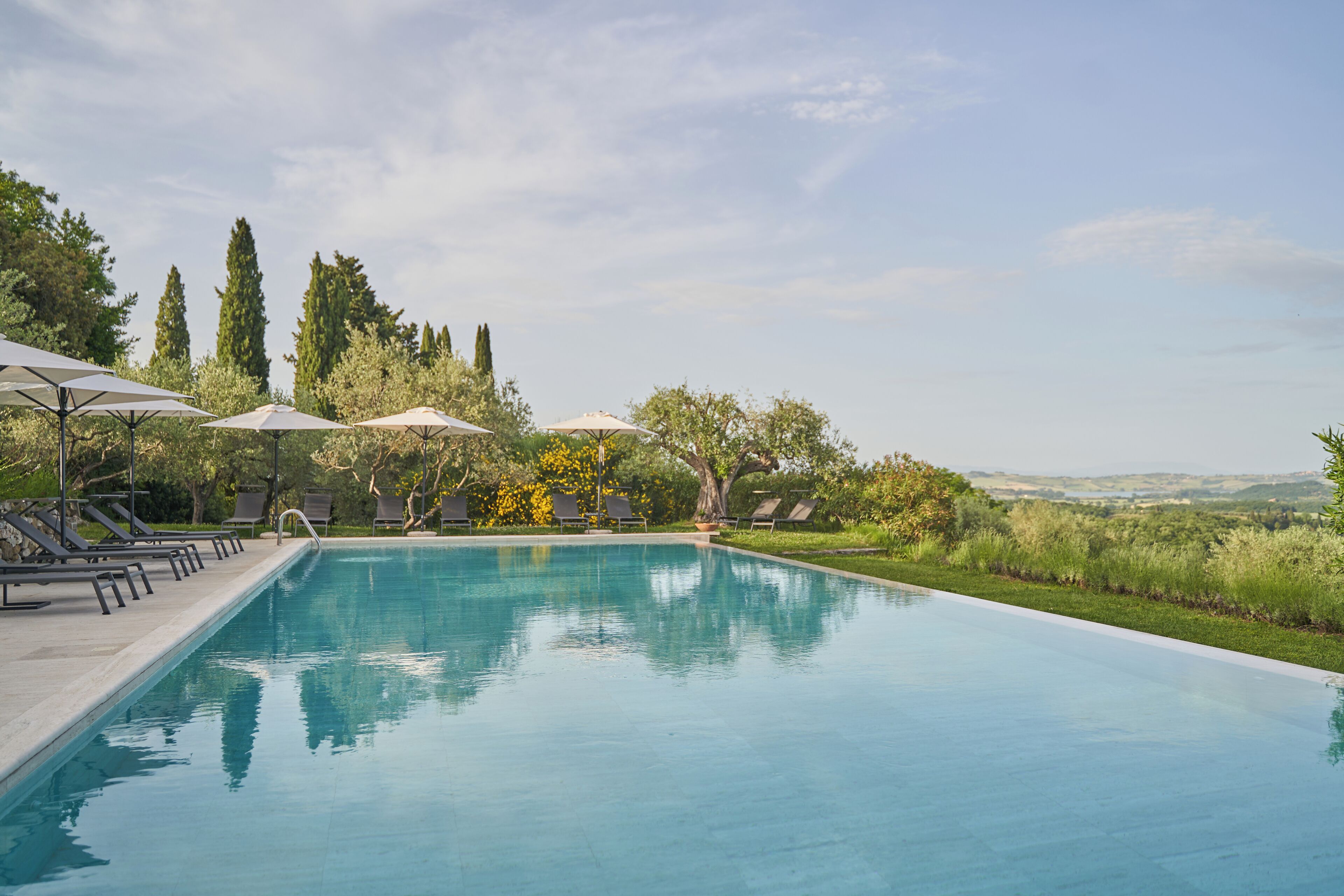 A swimming pool with lounge chairs and umbrellas, surrounded by trees and a view of distant hills.