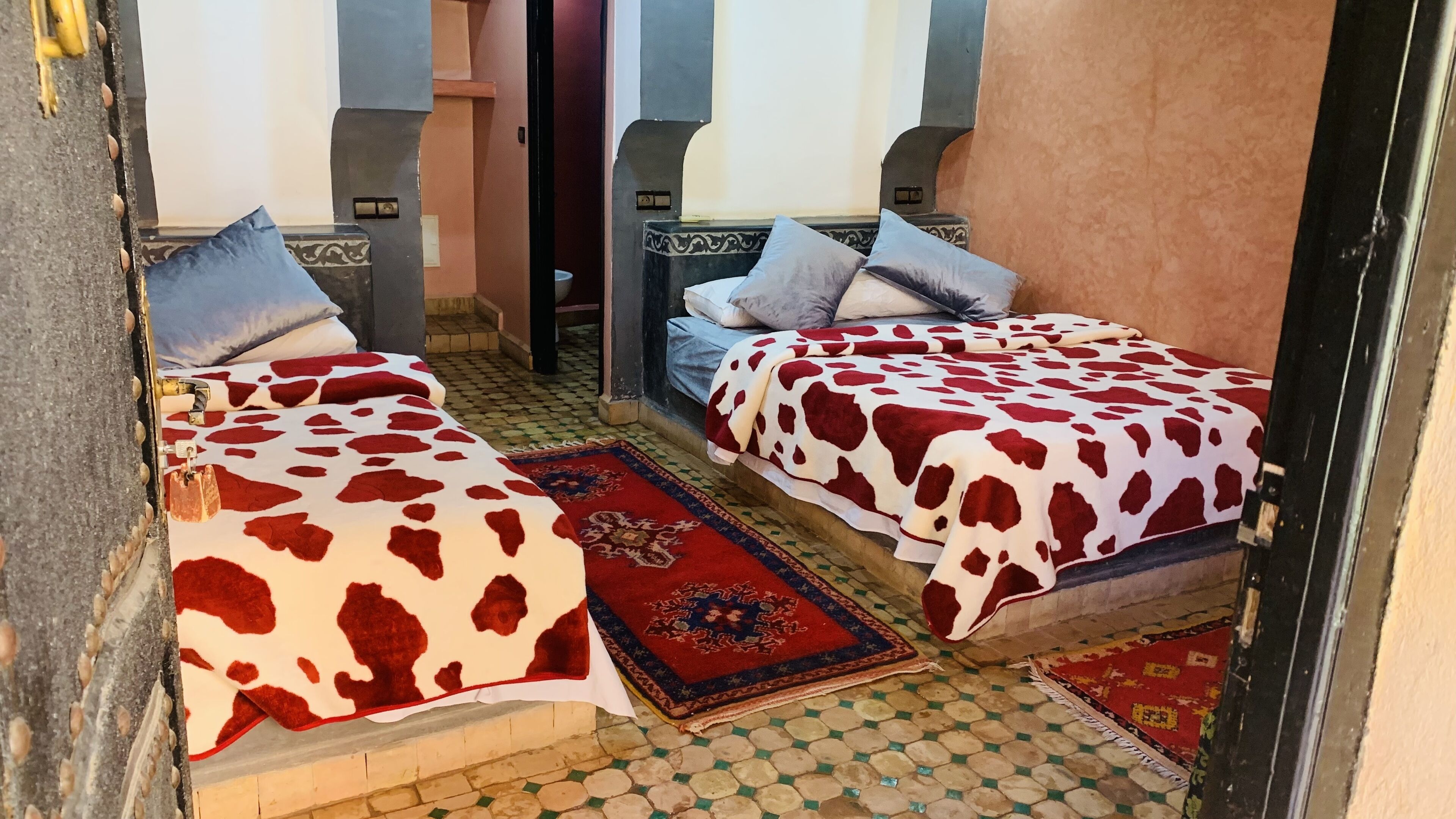 Una habitación tradicional con dos camas, cada una con una colcha estampada, un tapete con motivos azules y rojos, y suelo de baldosas.