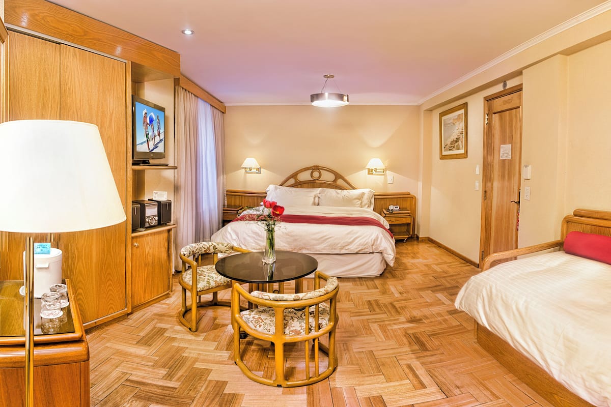 A hotel room with a bed, a desk with a lamp, a chair, a wooden wardrobe, and a television.