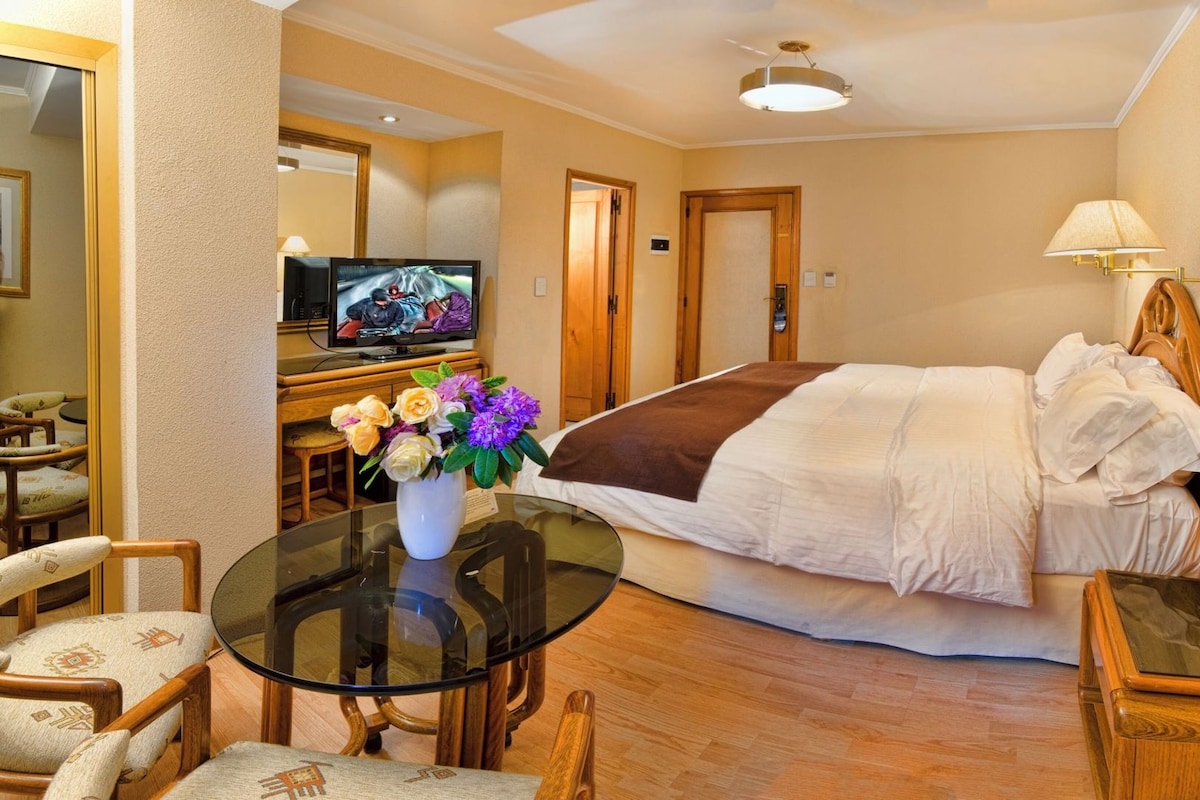 A hotel room with a bed, a TV, a table with chairs, and a vase of flowers.