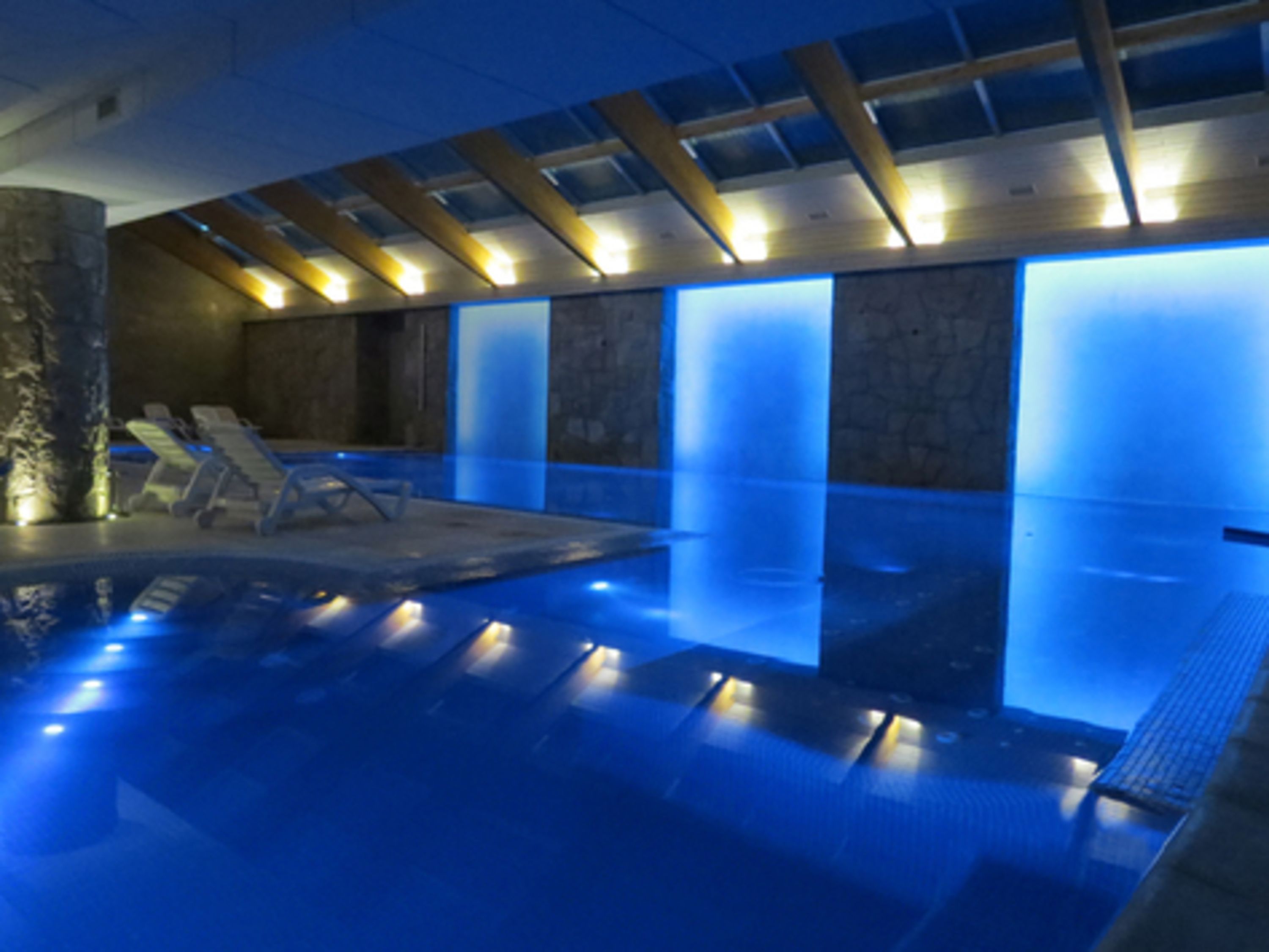 Een modern binnenzwembad met blauwe verlichting, lounge fauteuils en een stenen muur.