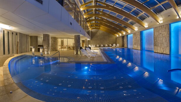 An indoor swimming pool with a curved design, a stone wall on one side, and a seating area with lounge chairs.