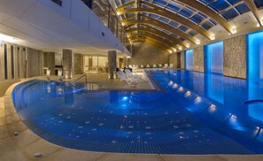 An indoor swimming pool with a curved design, a stone wall on one side, and a seating area with lounge chairs. - Hotel Cristal (San Carlos de Bariloche)