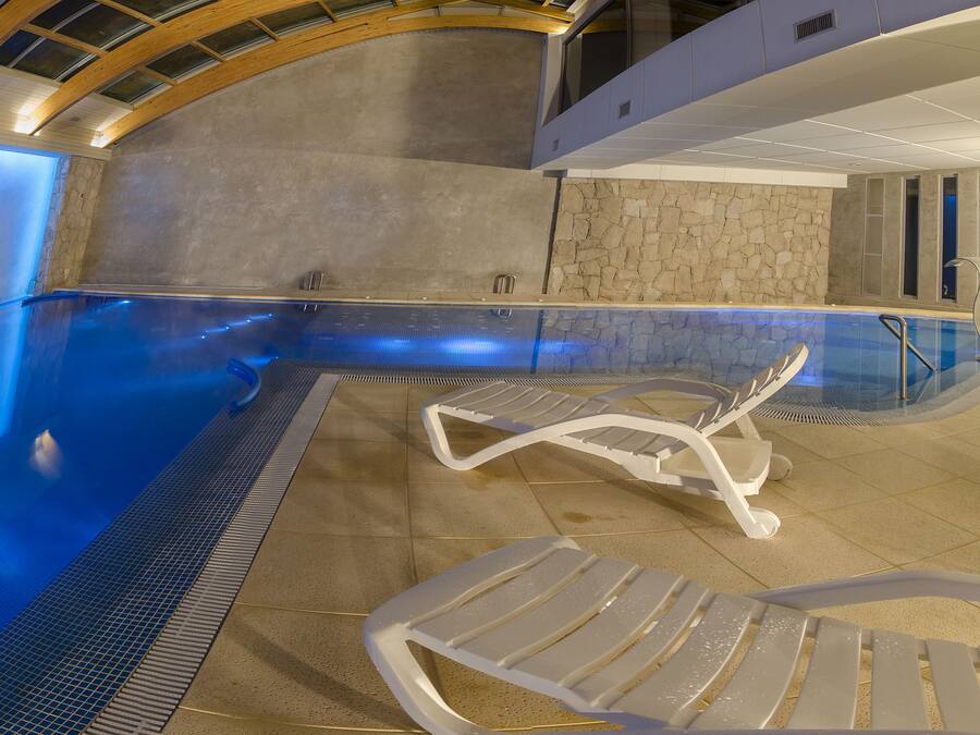 Une piscine intérieure moderne dotée d’un plafond incurvé, de murs en pierre et d’un espace détente avec des chaises longues blanches.