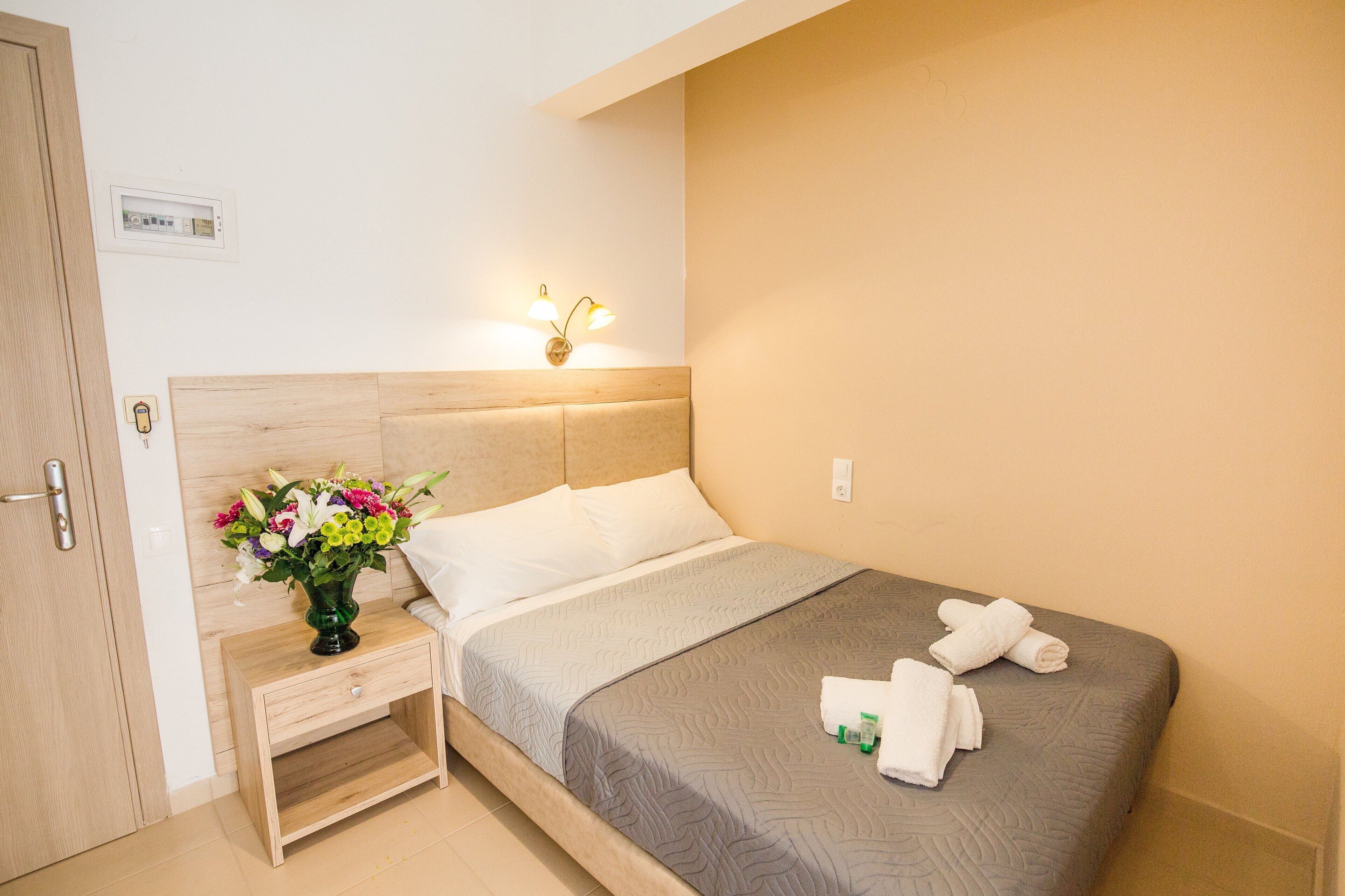 A neatly made bed with a grey quilt, a bedside table with a vase of flowers, and a wall-mounted light fixture.