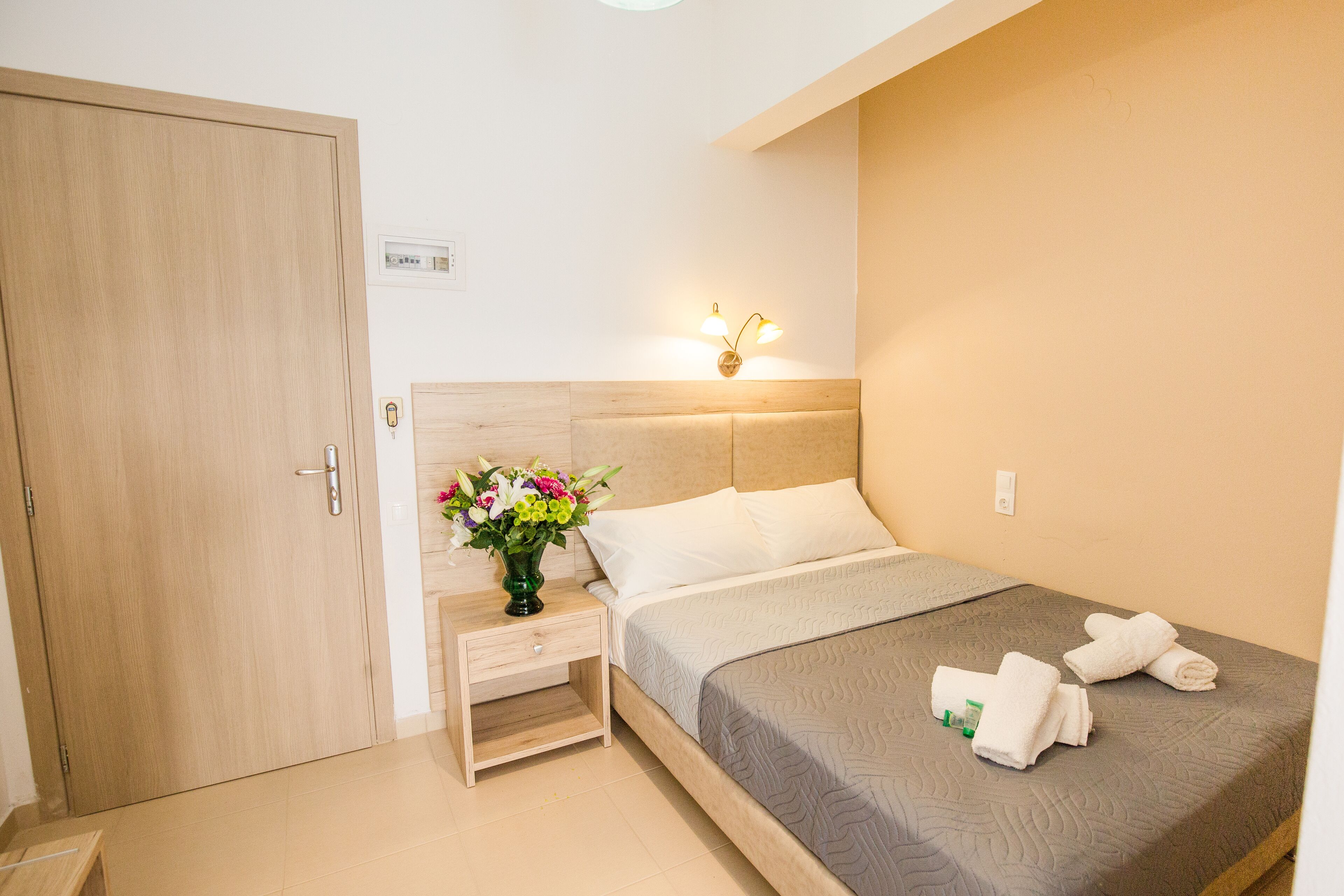 A hotel room with a wooden door, a bedside table with a vase of flowers, a bed with a gray quilt, and a wall-mounted light fixture.