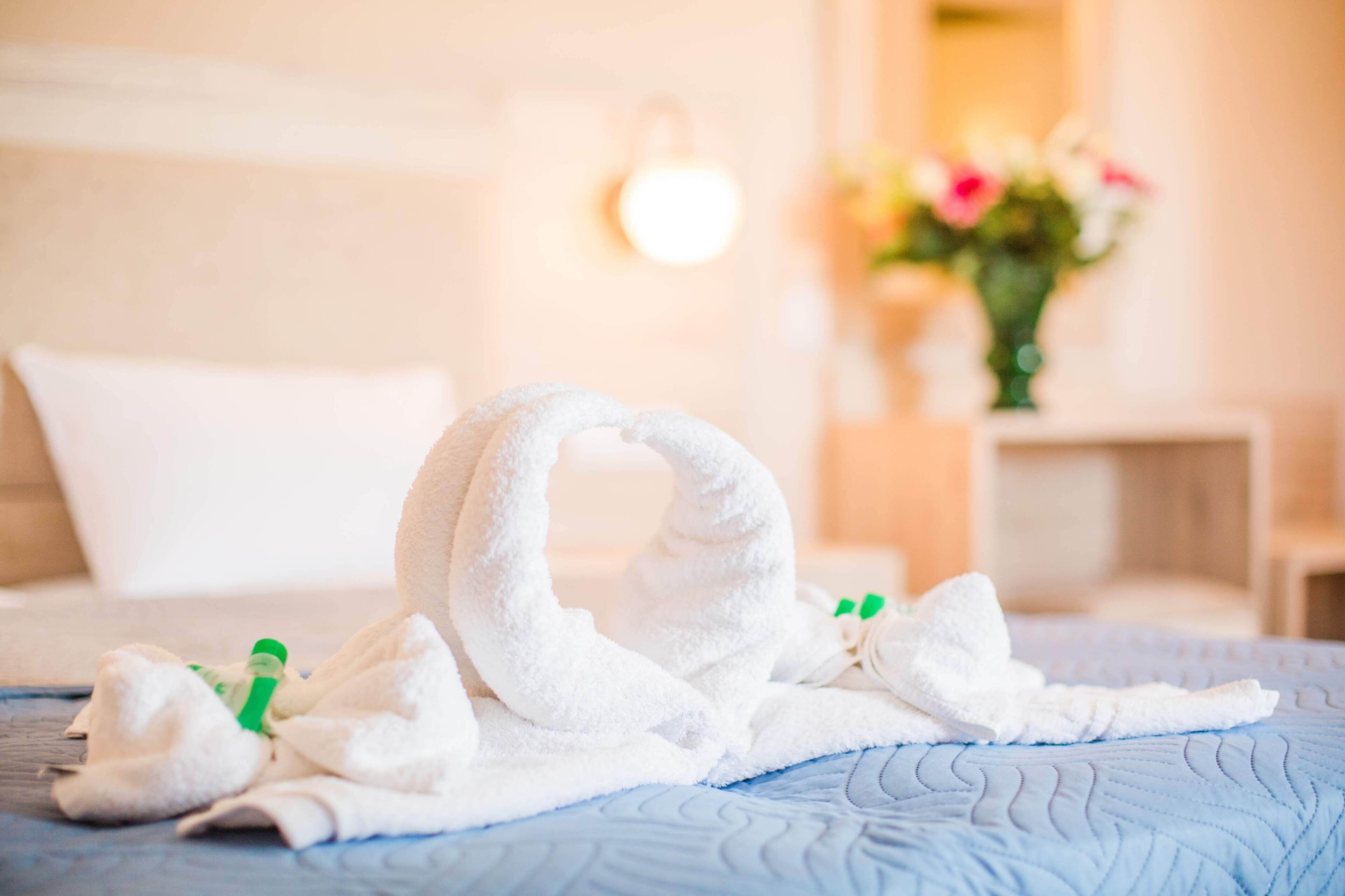 Una cama bien hecha con un edredón azul, una toalla blanca doblada en forma de cisne y una mesita de noche con una botella verde y un objeto blanco.